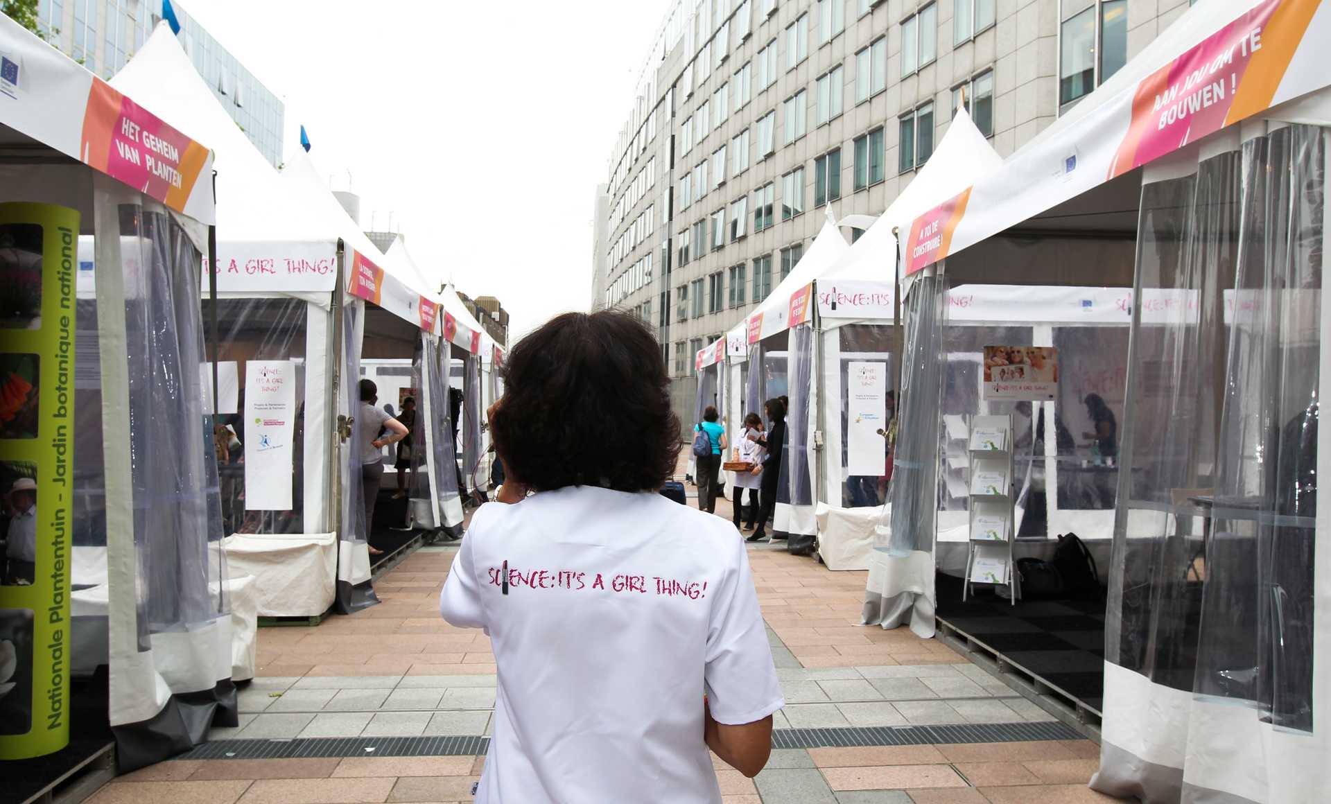 A person with shoulder-length hair is seen walking through a series of outdoor conference booths. Red writing on the back of their lab coat reads: Science: its a girl thing!