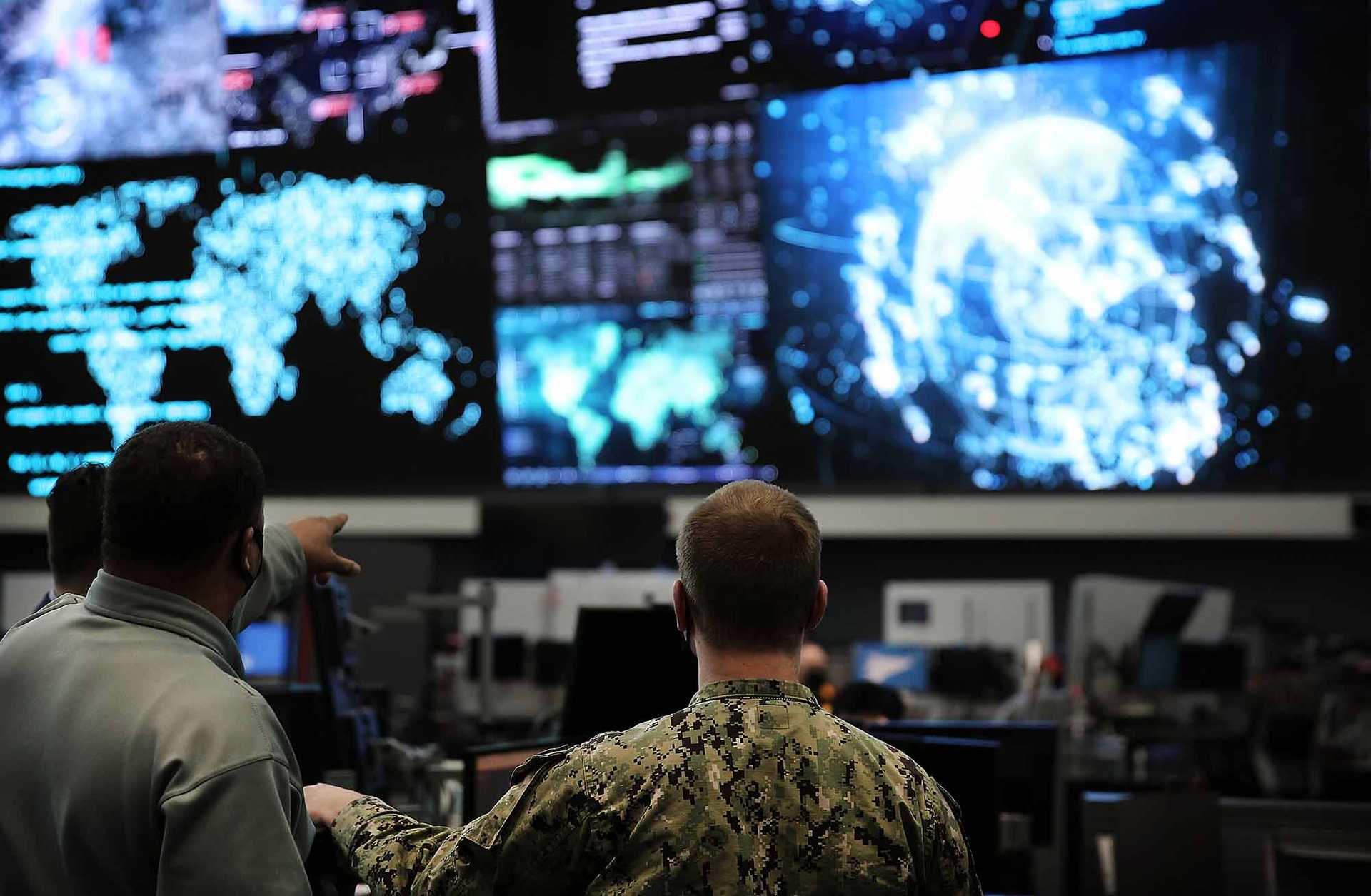 Two men in military uniform are seen from behind, looking up at large screens showing world maps, graphs, and other information.