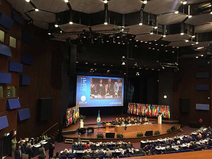 Several people gather in a large conference room. On stage is a high table as well as diverse flags and a huge display for presentations.