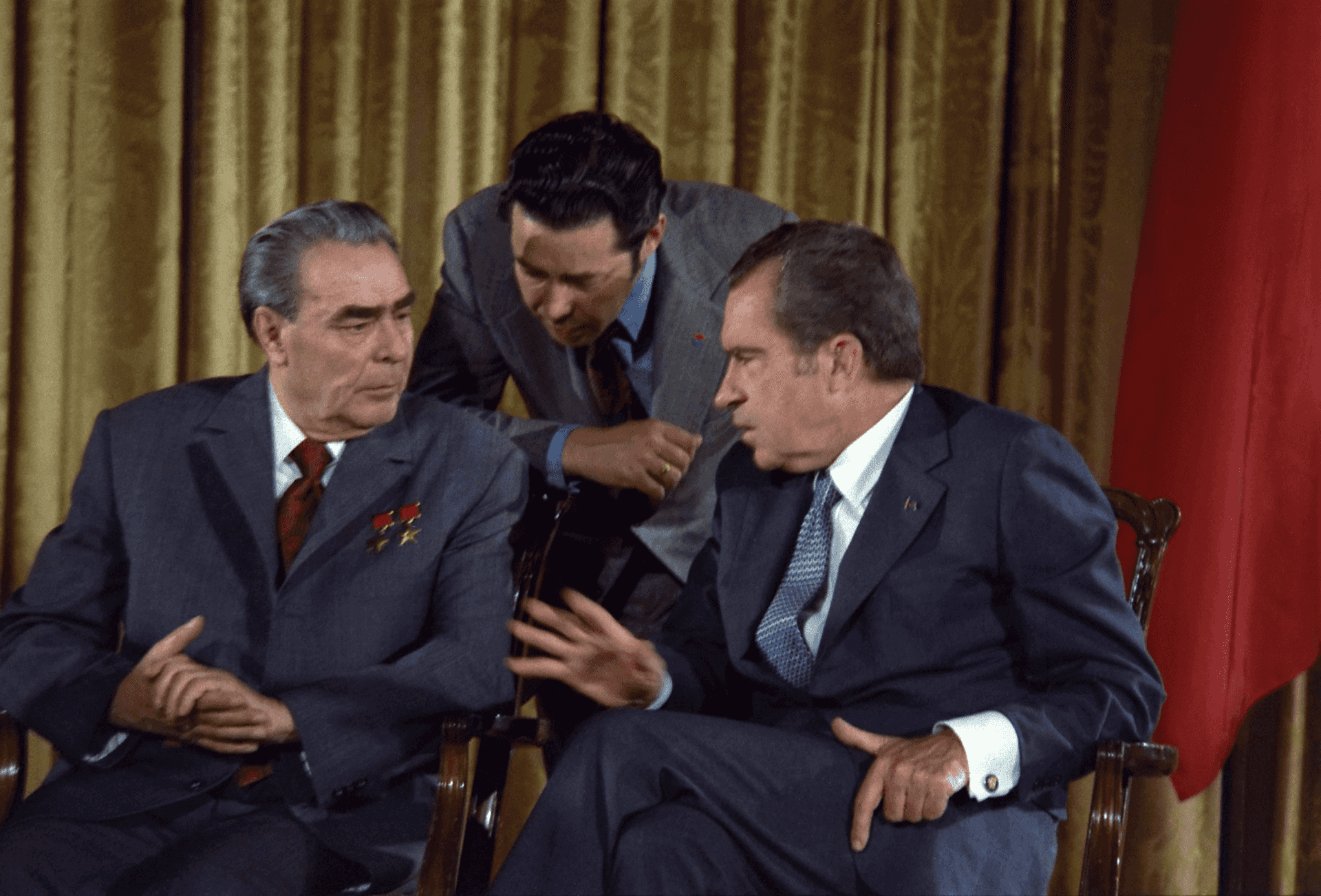 Nixon and Brezhnev sit together, seemingly on a boat or a terrace by the water. They gesture; Brezhnev raising his index finger and Nixon pointing his thumb up.