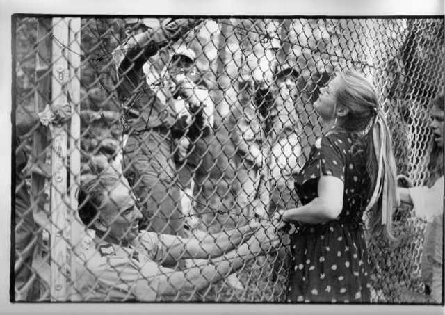 Black and white photograph shows a young blonde, white women facing a fence. She put her hands through it. On the other side of the fence there are men trying to push her away.